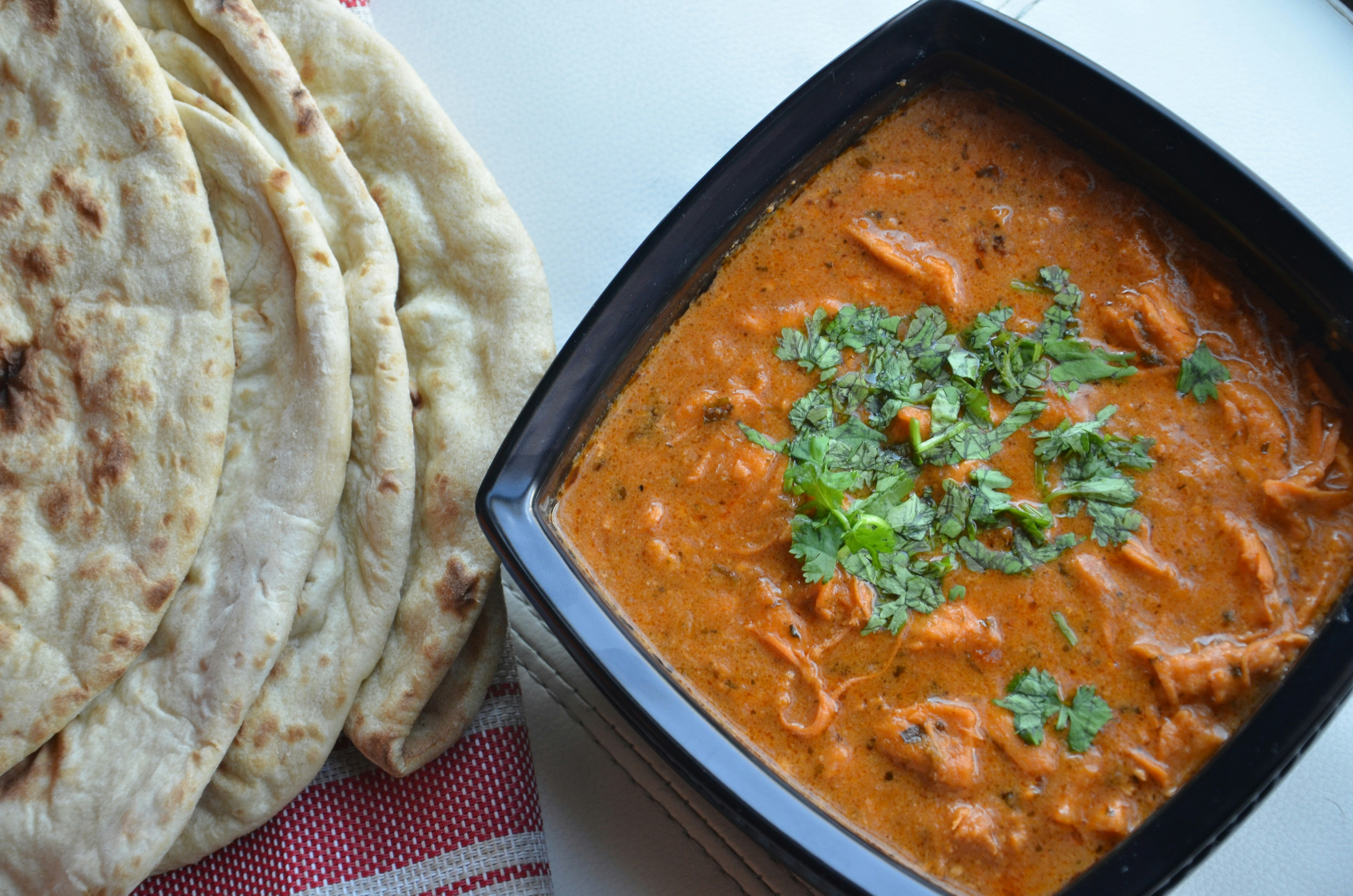 Creamy orange butter chicken in a dark bowl with fresh naan bread and cilantro garnish, warm Indian kitchen setting