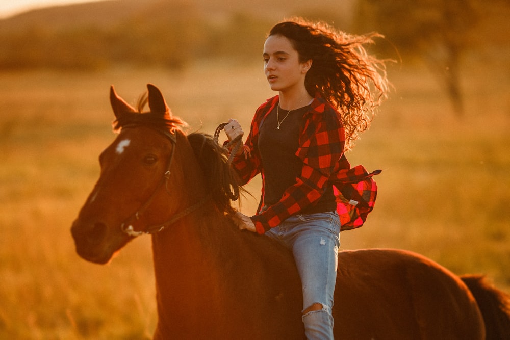 girl in red and black plaid dress shirt and blue denim jeans riding brown horse during