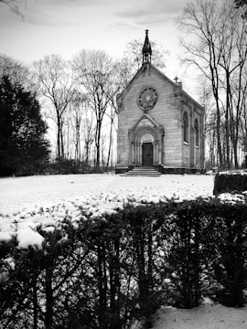 A small, old chapel with intricate architecture stands in a snow-covered landscape surrounded by bare trees. The building features a pointed roof with a small cross, arched windows, and a detailed circular window above the entrance. In the foreground, a hedge is partially covered in snow, adding texture to the scene.