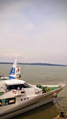 A large white ship with blue accents sits on a body of water. Several people in uniforms are visible on the deck of the vessel. The sky is overcast with shades of grey and there is a distant landmass across the water.