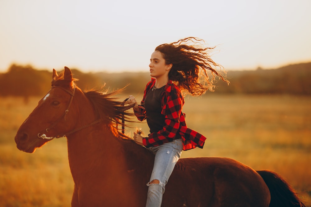 mujer con camisa de vestir a cuadros rojos y negros y jeans de mezclilla azules montando caballo marrón durante