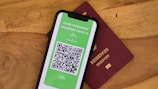 A smartphone displaying a digital COVID-19 passport with a QR code is placed on top of a maroon-colored physical passport. The digital document is green with text and a black and white QR code, while the physical passport has gold embossed lettering.