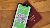 A smartphone displaying a digital COVID-19 passport with a QR code is placed on top of a maroon-colored physical passport. The digital document is green with text and a black and white QR code, while the physical passport has gold embossed lettering.