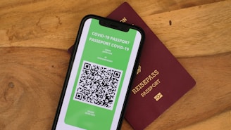A smartphone displaying a digital COVID-19 passport with a QR code is placed on top of a maroon-colored physical passport. The digital document is green with text and a black and white QR code, while the physical passport has gold embossed lettering.