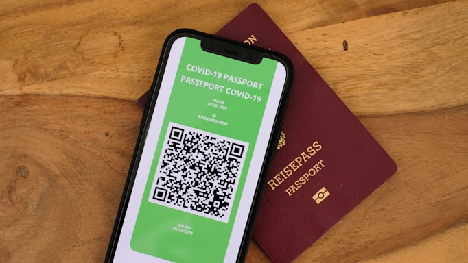 A smartphone displaying a digital COVID-19 passport with a QR code is placed on top of a maroon-colored physical passport. The digital document is green with text and a black and white QR code, while the physical passport has gold embossed lettering.
