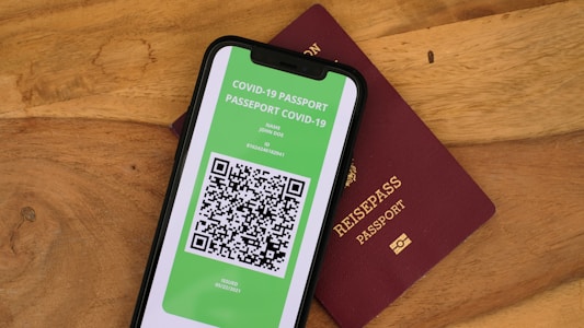A smartphone displaying a digital COVID-19 passport with a QR code is placed on top of a maroon-colored physical passport. The digital document is green with text and a black and white QR code, while the physical passport has gold embossed lettering.