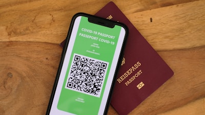 A smartphone displaying a digital COVID-19 passport with a QR code is placed on top of a maroon-colored physical passport. The digital document is green with text and a black and white QR code, while the physical passport has gold embossed lettering.