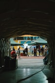 A tunnel leads out to a sports field where several people are gathered. The scene includes green tents, equipment, and crew members, suggesting a filming or event setup. The audience seats in blue and yellow are visible in the background, giving a stadium-like setting.