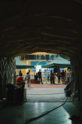 A tunnel leads out to a sports field where several people are gathered. The scene includes green tents, equipment, and crew members, suggesting a filming or event setup. The audience seats in blue and yellow are visible in the background, giving a stadium-like setting.