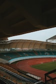 An empty sports stadium with a large, covered seating area and a red running track surrounding a green field. The stadium has teal and orange seating, and the architecture includes a sweeping canopy roof.