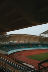 An empty sports stadium with a large, covered seating area and a red running track surrounding a green field. The stadium has teal and orange seating, and the architecture includes a sweeping canopy roof.