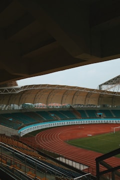 An empty sports stadium with a large, covered seating area and a red running track surrounding a green field. The stadium has teal and orange seating, and the architecture includes a sweeping canopy roof.