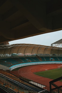 An empty sports stadium with a large, covered seating area and a red running track surrounding a green field. The stadium has teal and orange seating, and the architecture includes a sweeping canopy roof.