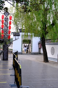 A serene scene in a traditional Chinese setting, featuring a white stone gateway with green calligraphy and surrounded by lush willows and other trees. Red lanterns hang to the left, adding a bright accent to the verdant environment. A wooden pathway leads up to railings and barriers, guiding the way towards the archway. Two people walk towards the gate, adding a sense of motion.