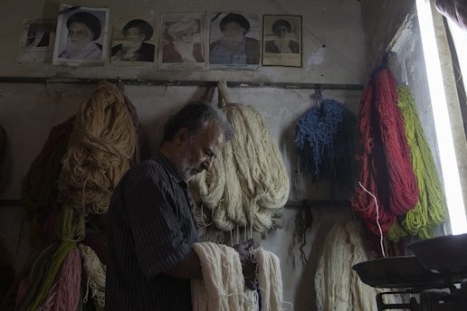 A man with gray hair and a beard stands in a room filled with bundles of colorful yarns hanging on the walls. The yarns are in various colors including red, blue, green, and beige. The room has a dimly lit atmosphere with natural light coming from a window. Above the yarns, there are several framed photographs of various individuals displayed on the wall.