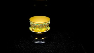 A beautifully ornate cup sits alone against a dark background. The cup features intricate patterns and decorations in gold and shades of green, adding to its luxurious appearance. The lighting highlights the decorative elements, creating a vivid contrast with the shadowy backdrop.
