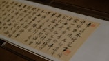 A piece of traditional East Asian calligraphy in black ink on a light beige paper, featuring neatly arranged characters in vertical columns. The writing is enclosed in a glass frame, and there are red seals stamped near the edges.