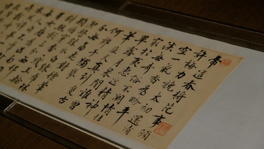 A piece of traditional East Asian calligraphy in black ink on a light beige paper, featuring neatly arranged characters in vertical columns. The writing is enclosed in a glass frame, and there are red seals stamped near the edges.