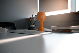 A serene setting featuring a desk in soft natural light. An orange portable fan is prominently placed beside a black mesh pen holder containing pencils. There are also a few notebooks stacked to the side.