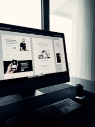 A sleek, modern workspace featuring a large computer monitor displaying a web design project. The screen shows multiple sections of a website with various texts and images. The setup includes a keyboard and mouse on a clean, dark surface with a small potted plant nearby. Natural light filters in through a window with sheer curtains, creating a soft ambiance.