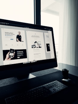 A sleek, modern workspace featuring a large computer monitor displaying a web design project. The screen shows multiple sections of a website with various texts and images. The setup includes a keyboard and mouse on a clean, dark surface with a small potted plant nearby. Natural light filters in through a window with sheer curtains, creating a soft ambiance.