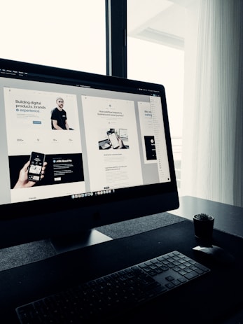 A sleek, modern workspace featuring a large computer monitor displaying a web design project. The screen shows multiple sections of a website with various texts and images. The setup includes a keyboard and mouse on a clean, dark surface with a small potted plant nearby. Natural light filters in through a window with sheer curtains, creating a soft ambiance.