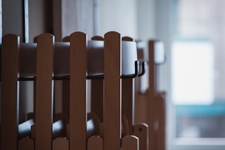 Wooden chairs are stacked in an orderly manner inside a room. Natural light filters through a window in the background, creating a calm and serene atmosphere.
