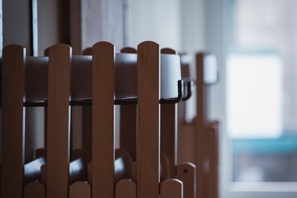 Wooden chairs are stacked in an orderly manner inside a room. Natural light filters through a window in the background, creating a calm and serene atmosphere.