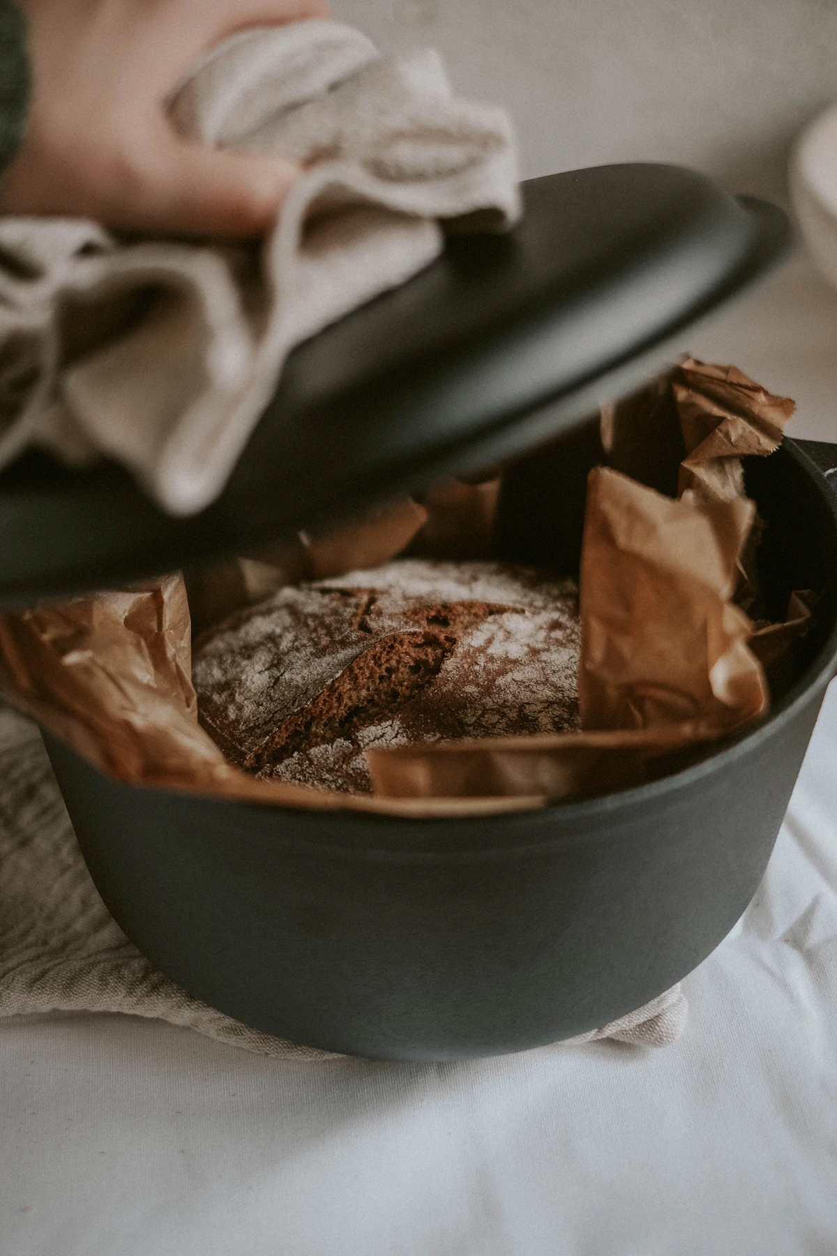 Hand with a linen cloth lifting the lid off a black Dutch oven of fresh bread
