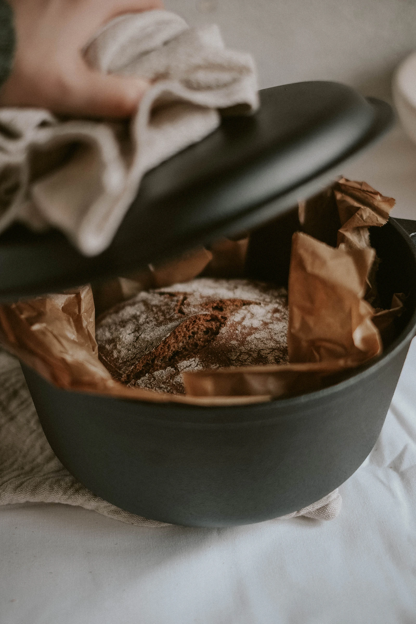 Hand with a linen cloth lifting the lid off a black Dutch oven of fresh bread