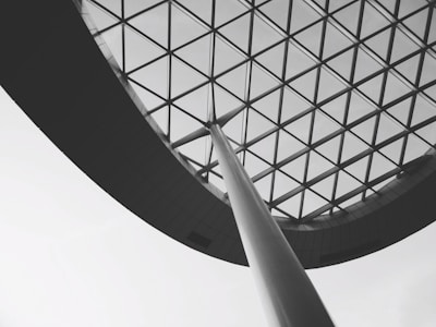 A modern architectural structure features a large, curved steel beam supporting an expansive glass ceiling with a geometric grid pattern. The image is in black and white, emphasizing the contrast between the linear elements and the open sky beyond.