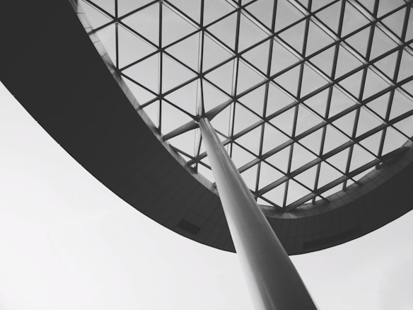 A modern architectural structure features a large, curved steel beam supporting an expansive glass ceiling with a geometric grid pattern. The image is in black and white, emphasizing the contrast between the linear elements and the open sky beyond.