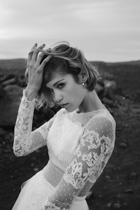 A person with short hair dressed in an intricate lace wedding dress is captured in a black and white photograph. The subject is holding their hair with one hand, standing in an outdoor environment with a somewhat barren and rocky background under a cloudy sky.