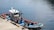 A small, colorful fishing boat is moored at a concrete pier. The boat has vivid blue and red accents and contains several pieces of equipment and ropes on its deck. The surrounding water is calm, with gentle ripples reflecting light. A cylindrical object and various nautical tools are visible on the boat's roof.
