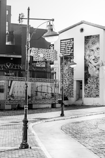 A black and white street scene featuring an industrial-style streetlamp, a building with a large mural of a smiling person, and a sign with a quote encouraging curiosity and looking up at the stars. The area has cobblestone and tiled pavement, with some artistic elements and industrial structures in the background.