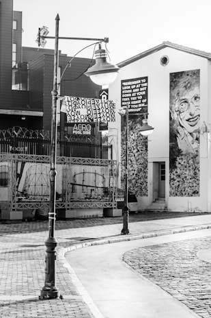 A black and white street scene featuring an industrial-style streetlamp, a building with a large mural of a smiling person, and a sign with a quote encouraging curiosity and looking up at the stars. The area has cobblestone and tiled pavement, with some artistic elements and industrial structures in the background.