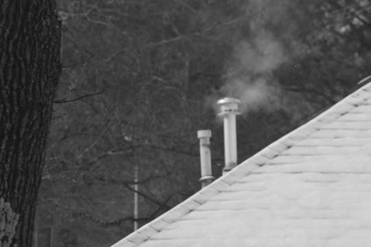 A snow-covered roof with two chimney pipes emitting smoke, set against a backdrop of bare winter trees.