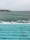 Swimmers are enjoying a swim in a large outdoor pool that overlooks the ocean. The pool's turquoise water contrasts with the darker, more turbulent sea beyond. Waves crash against the rocky shore, with a cluster of buildings visible on a distant headland.