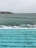 Swimmers are enjoying a swim in a large outdoor pool that overlooks the ocean. The pool's turquoise water contrasts with the darker, more turbulent sea beyond. Waves crash against the rocky shore, with a cluster of buildings visible on a distant headland.