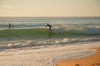 A surfer rides a wave near the shore at sunset, with the ocean stretching into the distance. The gentle waves create a rhythm along the sandy beach.