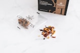 An open tea packet labeled 'Crystal Tea High Quality CBD' is on a white surface, with several dried tea leaves and fruit pieces spilling out next to it. In the background, there is a box labeled 'Elixir Immuno Acquamarina' with additional branding details.