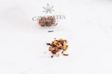 A thin plastic bag with a cannabis leaf logo and the text 'Crystal Tea High Quality CBD' contains a variety of dried tea leaves, scattered on a smooth white surface. The tea appears to consist of small pieces of dried fruit, leaves, and possibly herbs, with a mix of colors including red, brown, and orange.