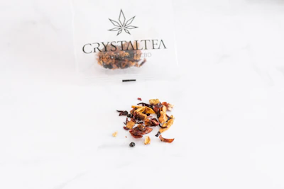 A thin plastic bag with a cannabis leaf logo and the text 'Crystal Tea High Quality CBD' contains a variety of dried tea leaves, scattered on a smooth white surface. The tea appears to consist of small pieces of dried fruit, leaves, and possibly herbs, with a mix of colors including red, brown, and orange.