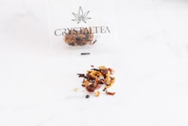 A thin plastic bag with a cannabis leaf logo and the text 'Crystal Tea High Quality CBD' contains a variety of dried tea leaves, scattered on a smooth white surface. The tea appears to consist of small pieces of dried fruit, leaves, and possibly herbs, with a mix of colors including red, brown, and orange.