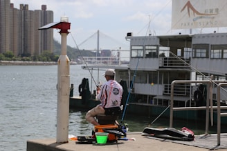 A person is seated on the edge of a pier, fishing beside a large boat marked 'Park Island'. In the background, a prominent bridge and high-rise buildings are visible across the water. Fishing equipment is spread out around the seated individual, who is wearing a cap and patterned shirt.