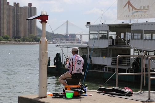 A person is seated on the edge of a pier, fishing beside a large boat marked 'Park Island'. In the background, a prominent bridge and high-rise buildings are visible across the water. Fishing equipment is spread out around the seated individual, who is wearing a cap and patterned shirt.