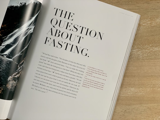 A magazine or book page is open on a wooden table. The title 'The Question About Fasting' is prominently displayed in large, bold fonts. Below the title, there is text in a smaller size. The page on the left shows part of an image featuring a waterfall, flowing over rocks surrounded by greenery.