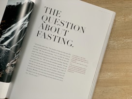 A magazine or book page is open on a wooden table. The title 'The Question About Fasting' is prominently displayed in large, bold fonts. Below the title, there is text in a smaller size. The page on the left shows part of an image featuring a waterfall, flowing over rocks surrounded by greenery.