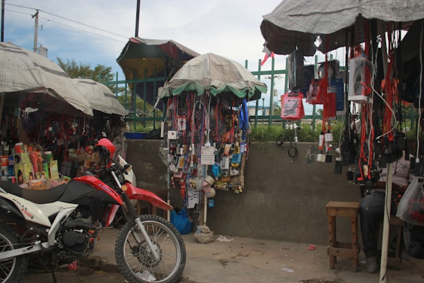 A busy street market stall with a variety of items hanging from the canopy-covered stands. A red and white motorcycle is parked in front of the stall, surrounded by various goods such as electronics, phone accessories, and personal care products. The stall is covered with makeshift umbrellas providing shade.