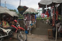 A busy street market stall with a variety of items hanging from the canopy-covered stands. A red and white motorcycle is parked in front of the stall, surrounded by various goods such as electronics, phone accessories, and personal care products. The stall is covered with makeshift umbrellas providing shade.